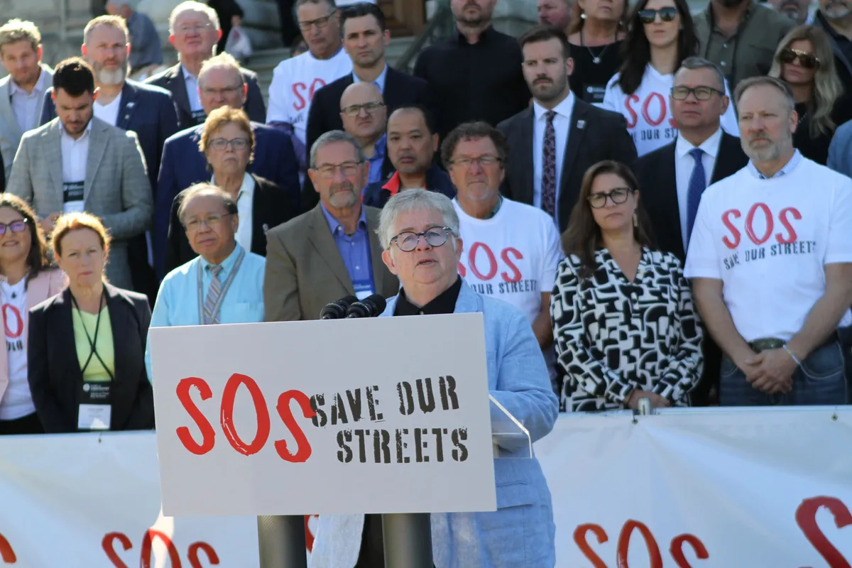 A speaker at the rally podium, with mayors and councillors standing behind, some in Save Our Streets T-shirts